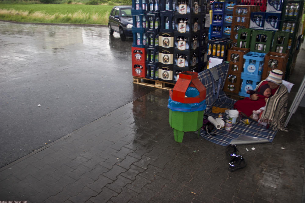 ﻿Krasser Regen. In der Mittagspause hat es uns voll erwischt. Lustigerweise genau bei dem Rewe, bei dem wir auch vor zwei Jahren Regenpause gemacht haben. Mit Klaris Picknickdecke bauen wir eine einigermaßen komfortable Schutznische.